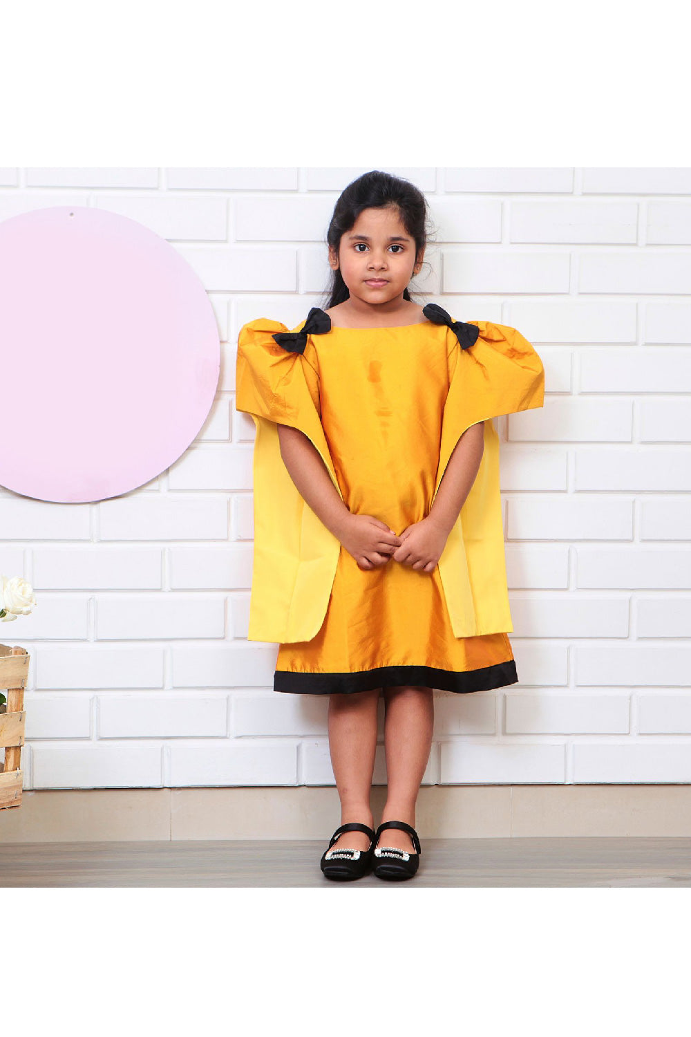 A young girl wearing a mustard yellow taffeta dress with black bow detailing and a round neckline.